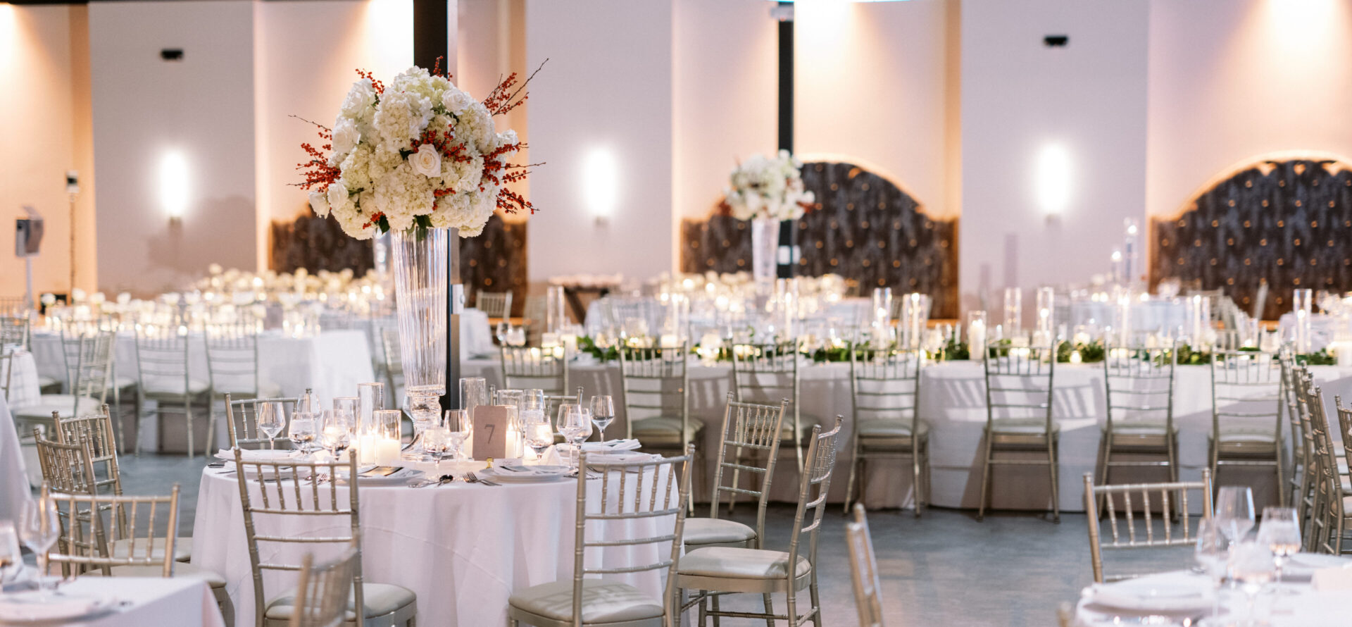 Elegant banquet hall with round tables, white linens, floral centerpieces, and gold chairs set for an event.