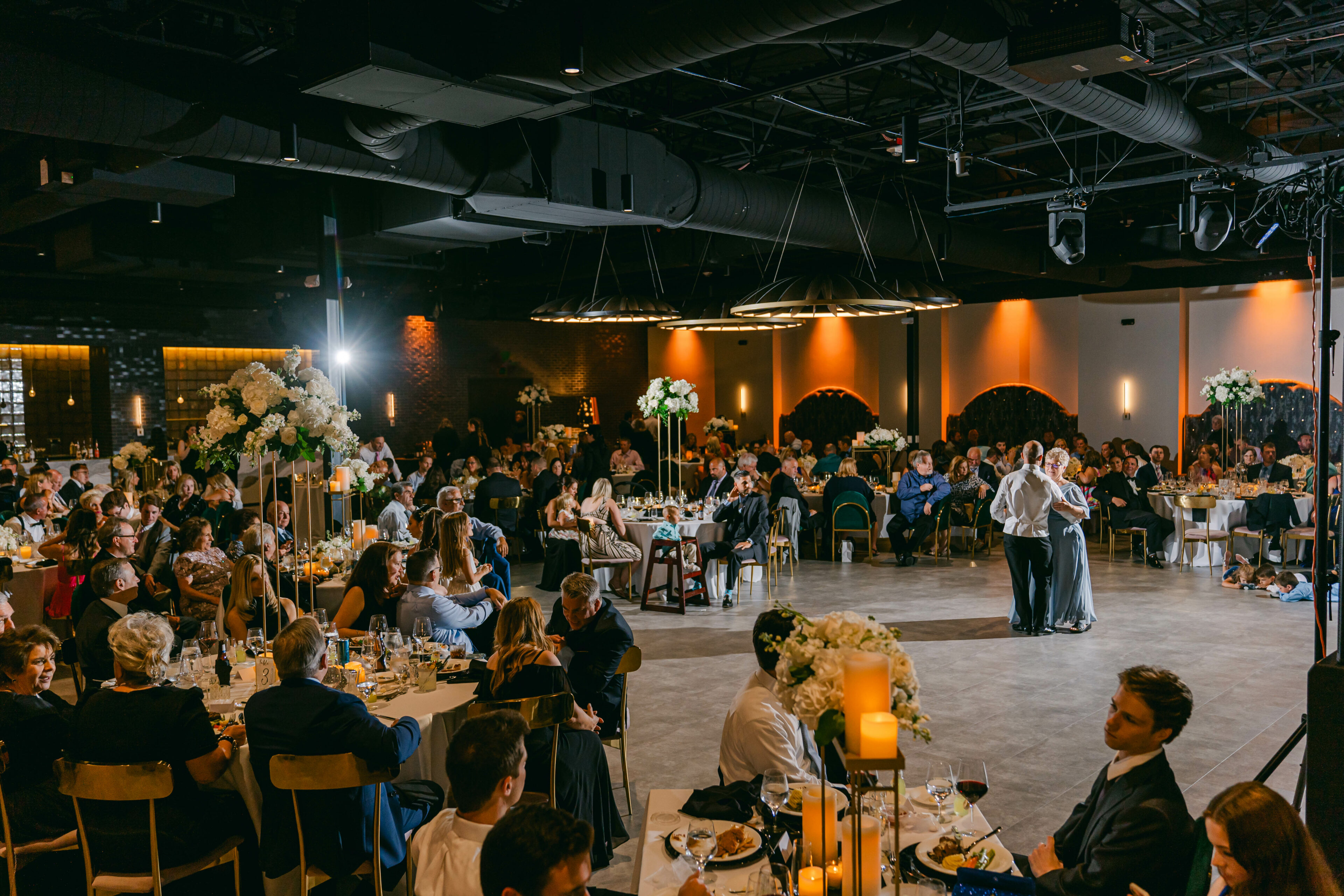 A couple shares their first dance at a wedding reception, surrounded by seated guests in a decorated hall.